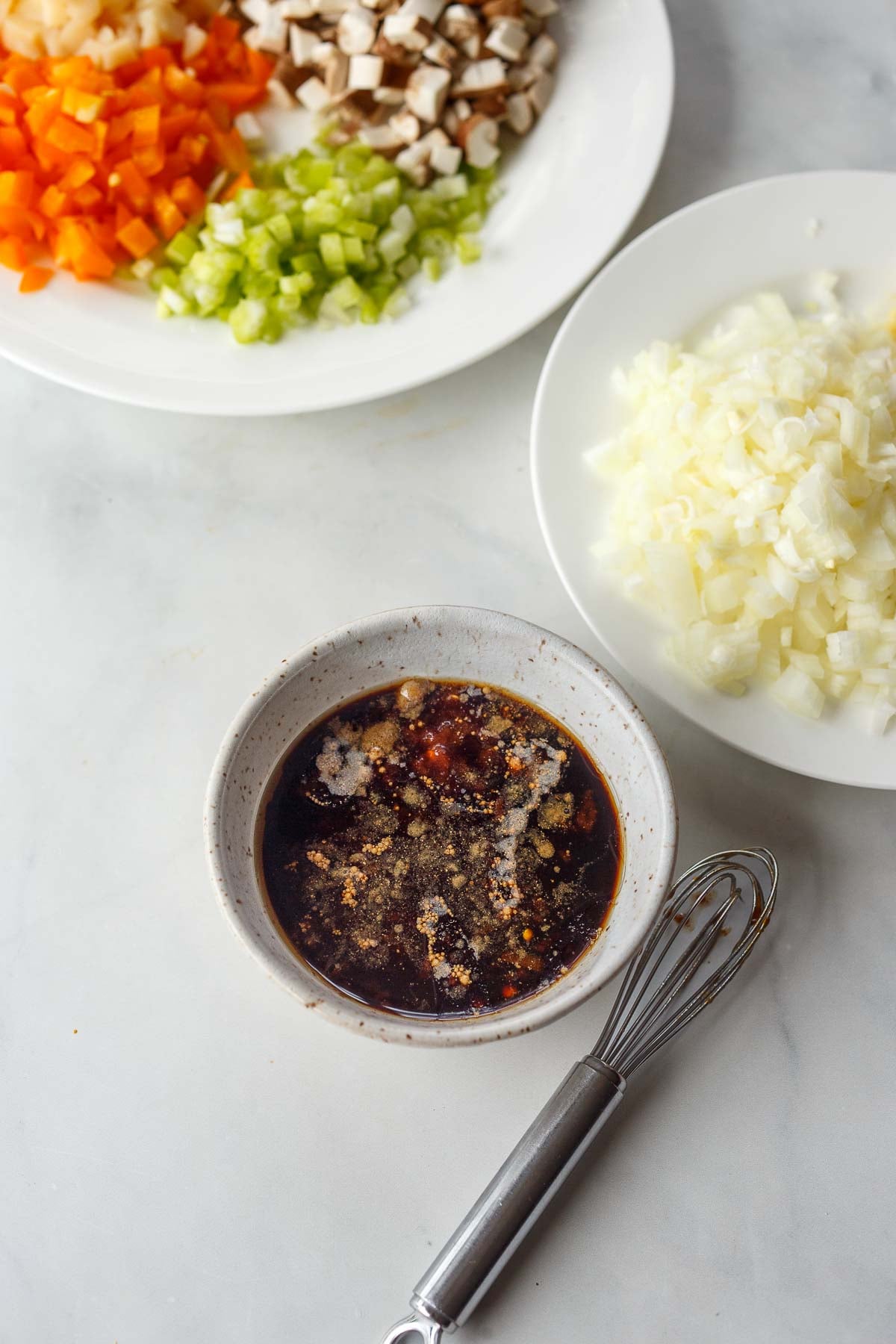 small bowl of Asian sauce with tiny whisk next to plates of veggies. 