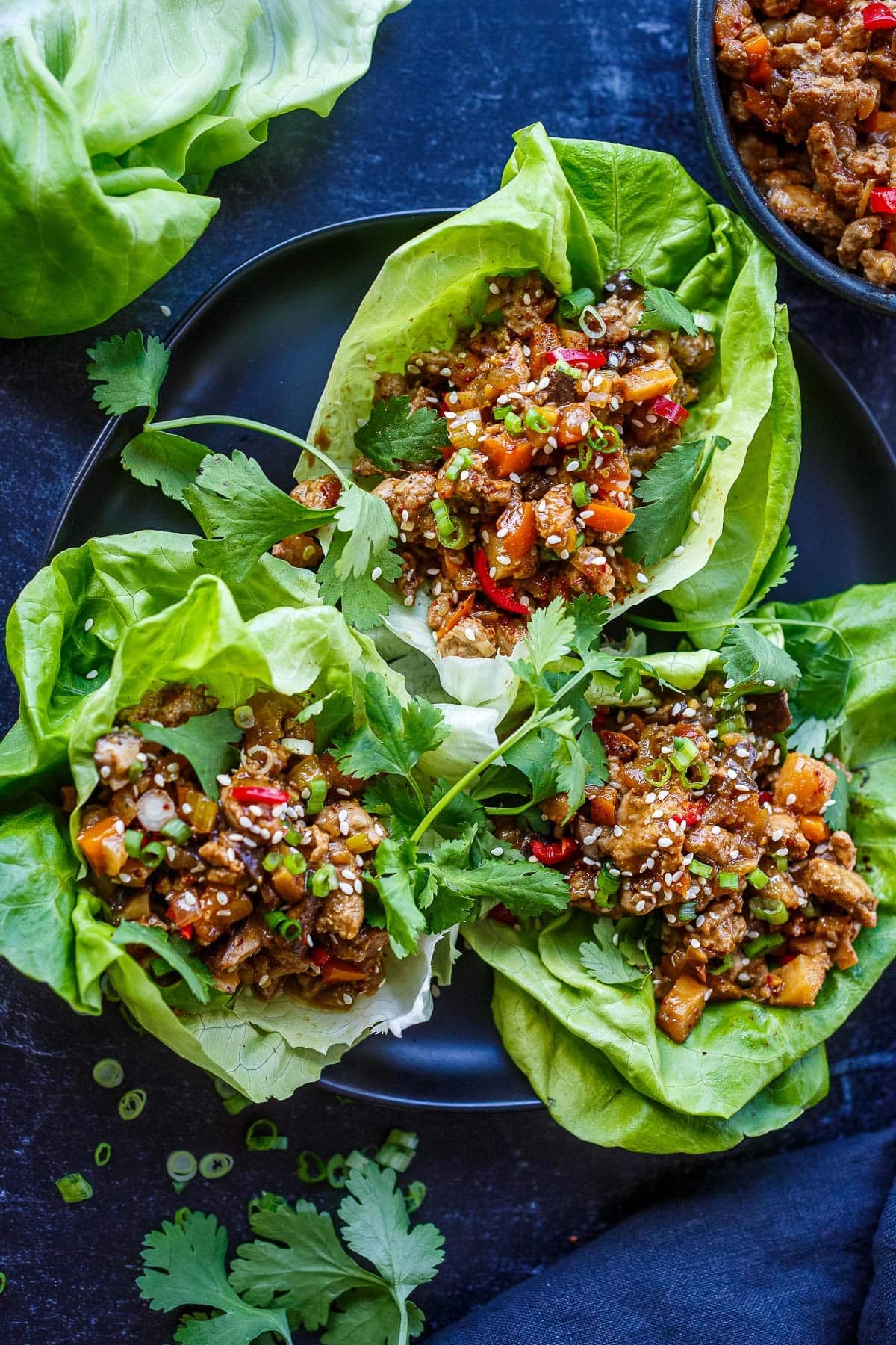 three Asian lettuce wraps on plate with saucy meat filling topped with fresh cilantro, green onions, and sesame seeds. 