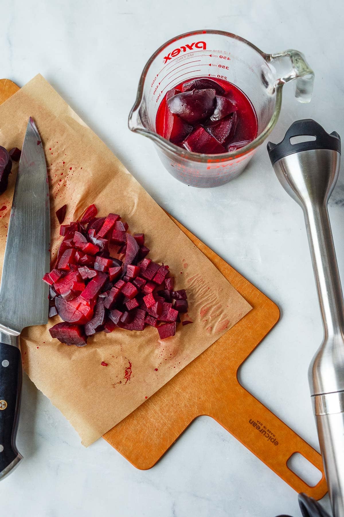 diced beets on a parchment-lined cutting board with sharp knife next to immersion blender and measuring cup with beets and broth.