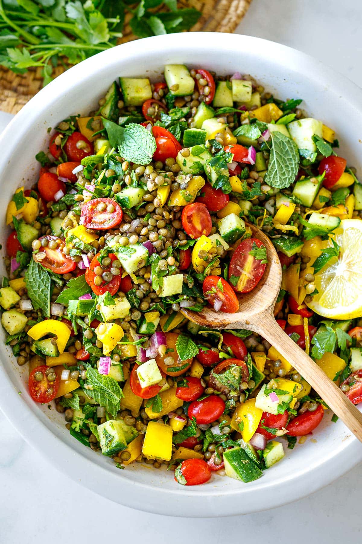 large bowl of Mediterranean Lentil Salad with peppers, cherry tomatoes, cucumber, fresh herbs, red onion, lemon half, and wood spoon for serving. 