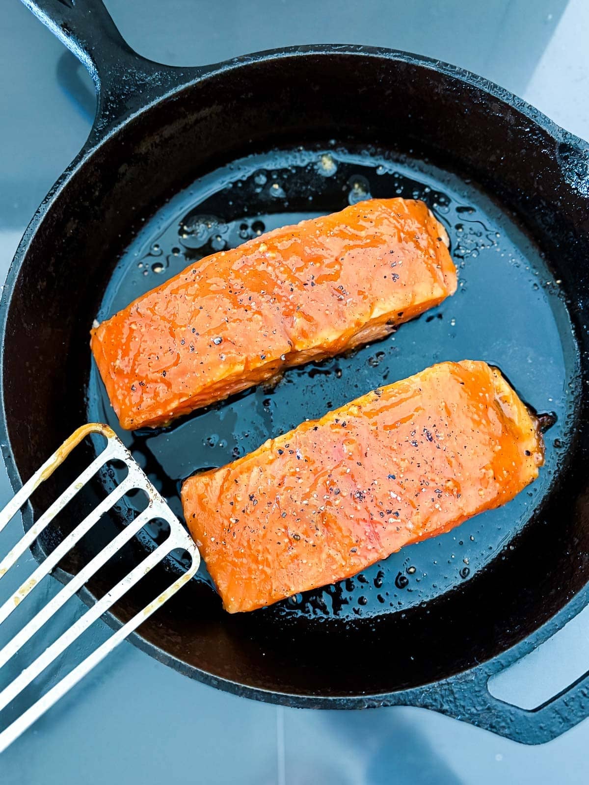 two salmon filets searing in a cast iron skillet garnished with black pepper - metal spatula resting on the side. 