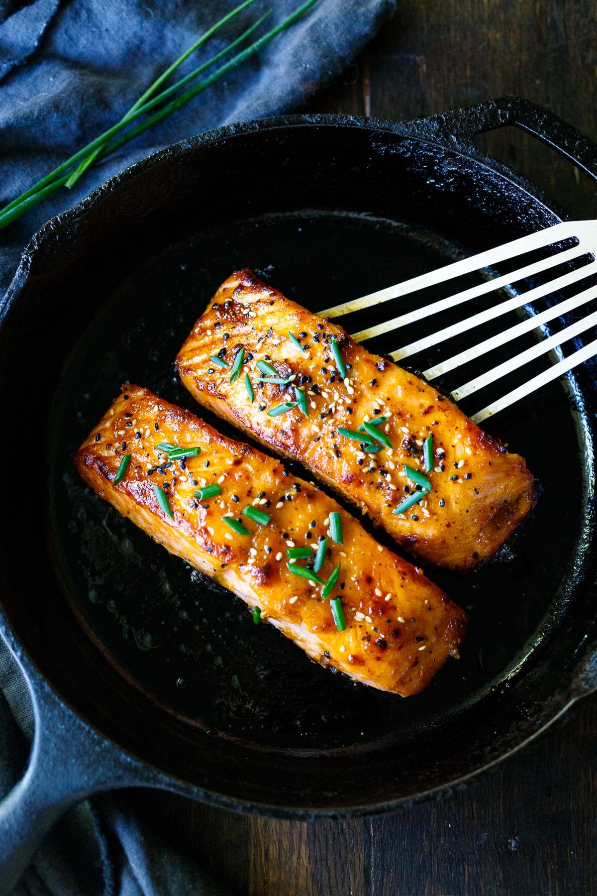 two broiled salmon filets on a cast iron skillet with sesame seeds and chives as garnish with a metal spatula beginning to lift one up.