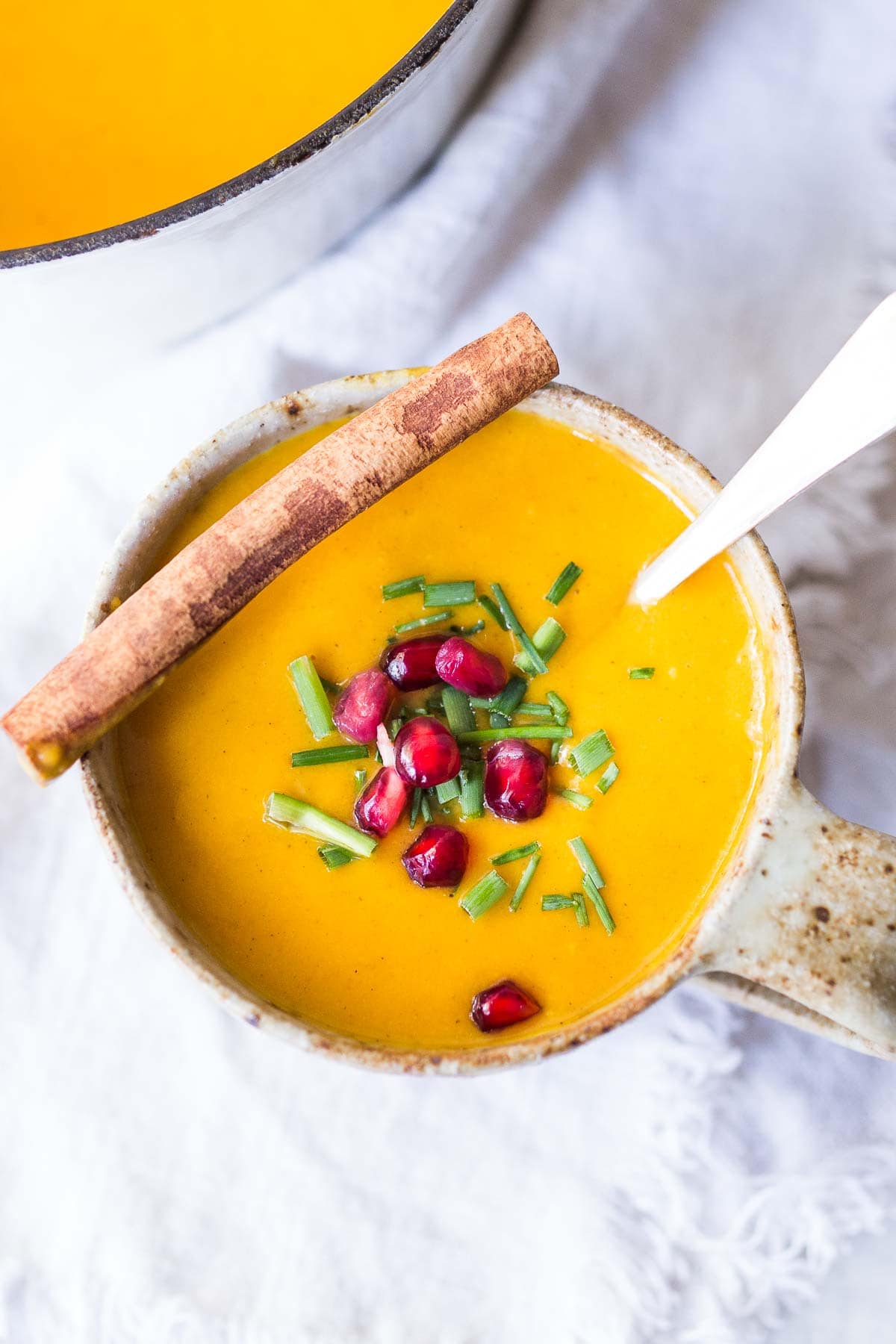 mug of Moroccan carrot soup topped with pomegranate seeds and chives with a cinnamon stick and spoon. 