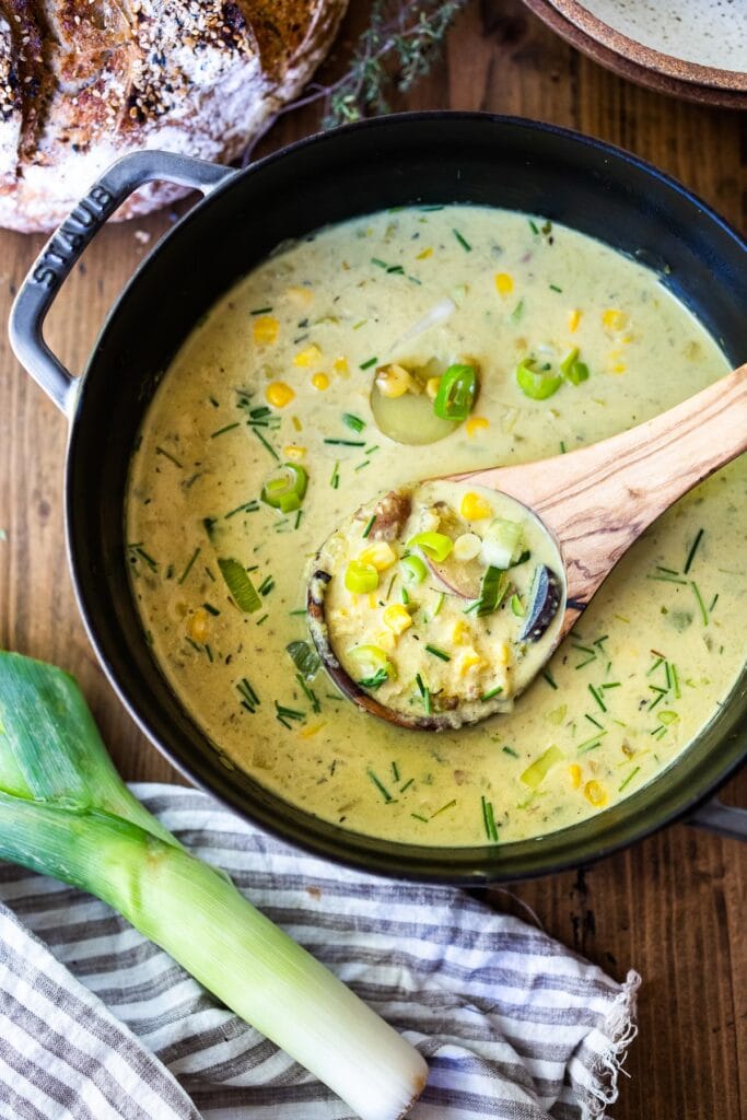 pot of coconut corn chowder with a wooden ladle lifting soup from the pot, showing corn, green onions, chives, and leeks.