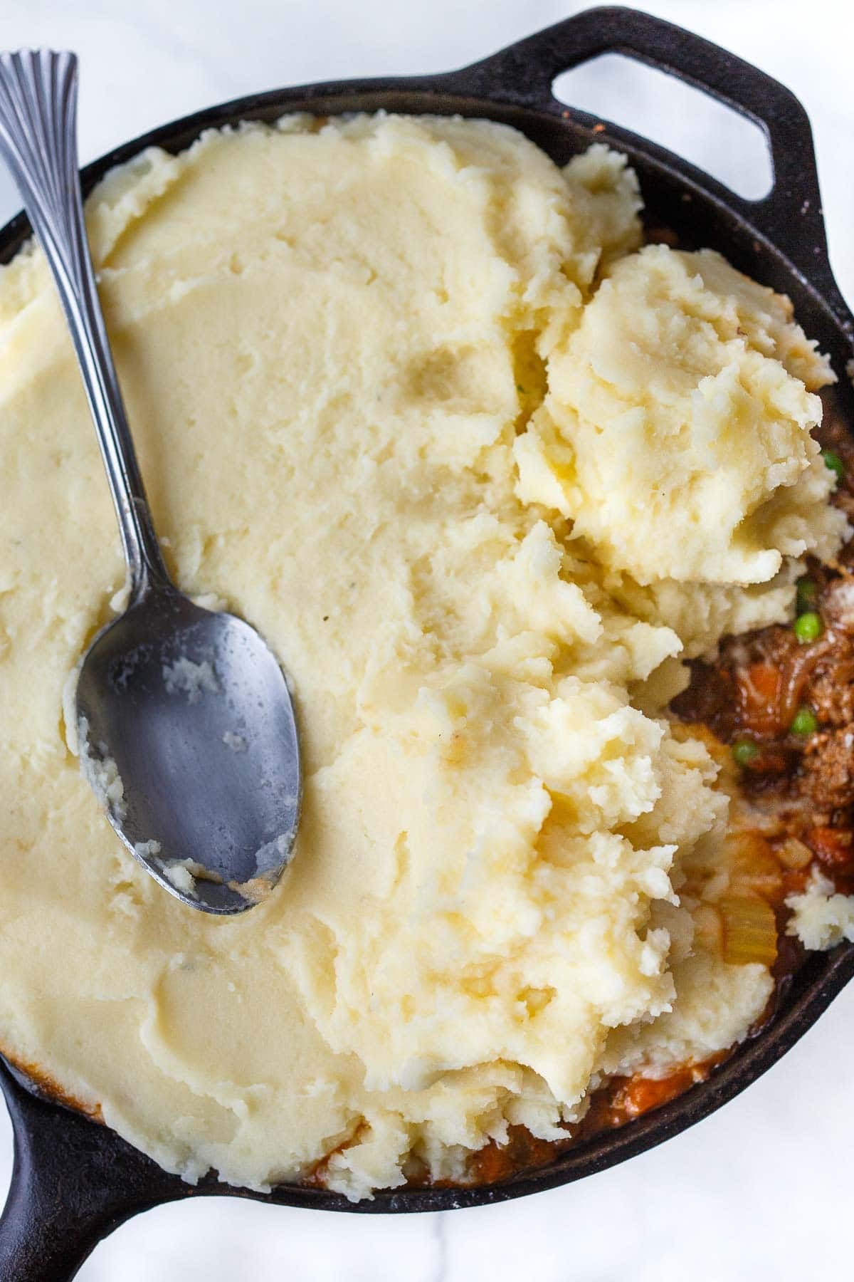 spoon spreading mashed potatoes over cottage pie filling in cast iron skillet.