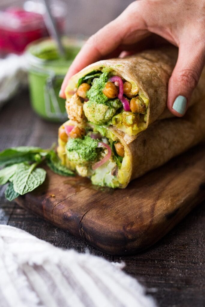 Best vegan meal: wood board with Frankies, Indian burritos, stacked on top, revealing an interior of cauliflower, curried potatoes, pickled onions, and mint chutney; hand holding the top of the burritos.