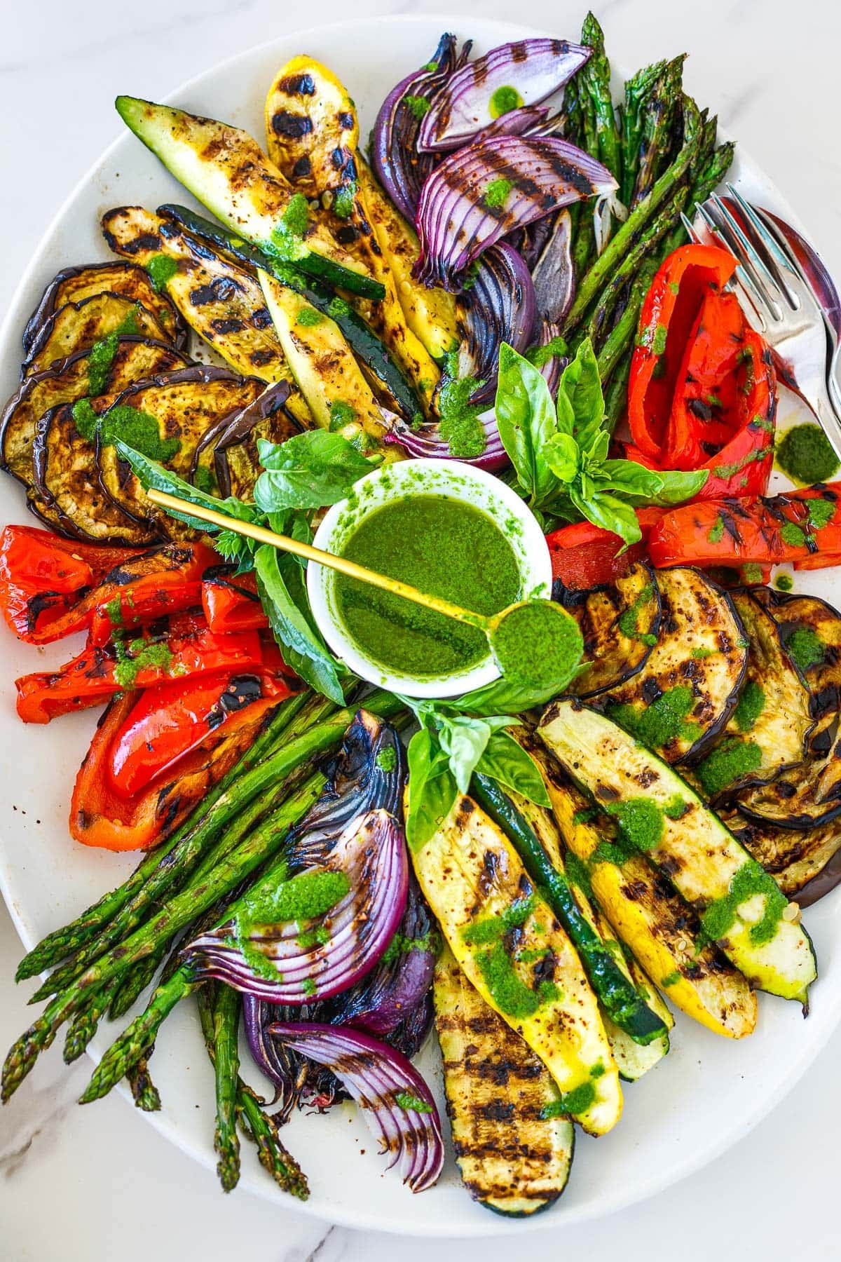 serving plate of grilled veggies surrounding a bowl of basil vinaigrette with gold spoon holding dressing on top.