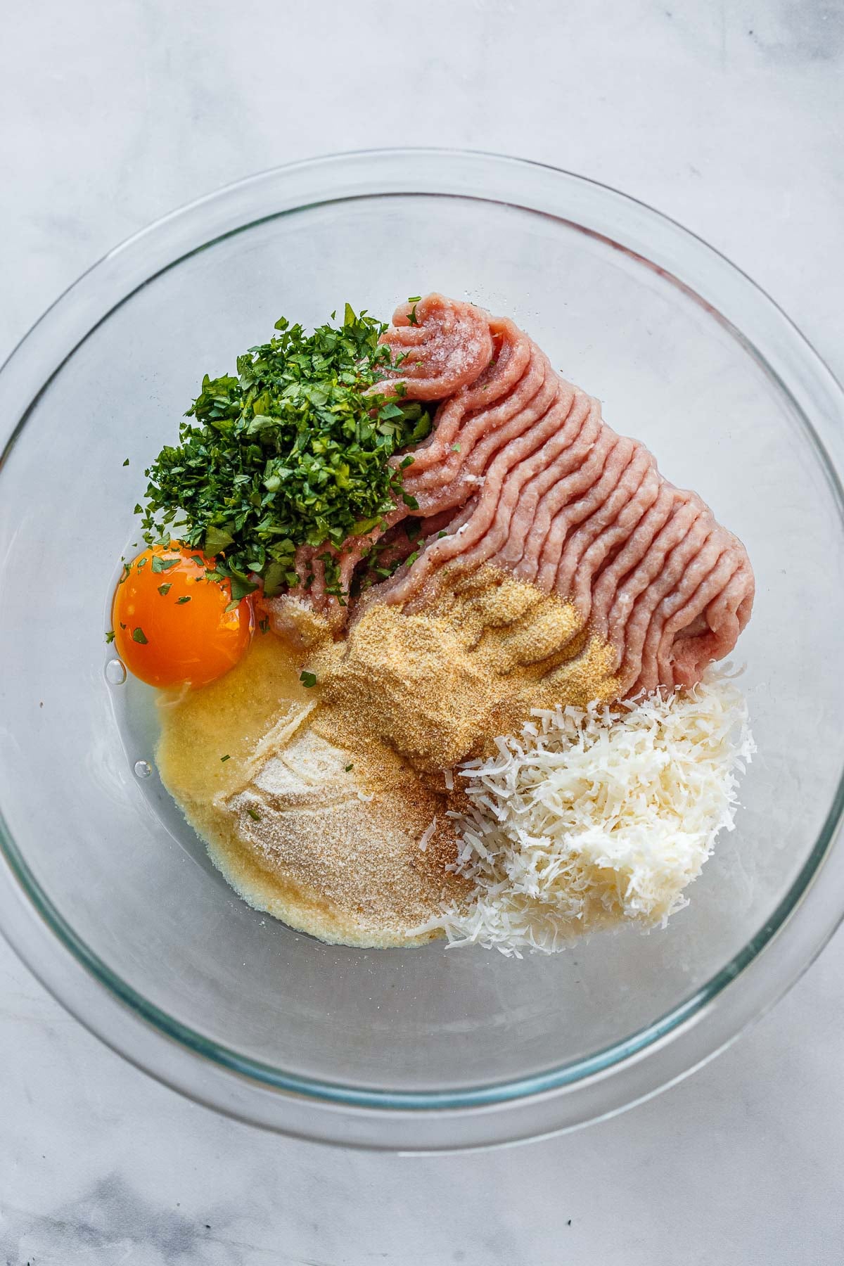 glass mixing bowl with ground meat, minced parsley, egg, spices, and parmesan. 