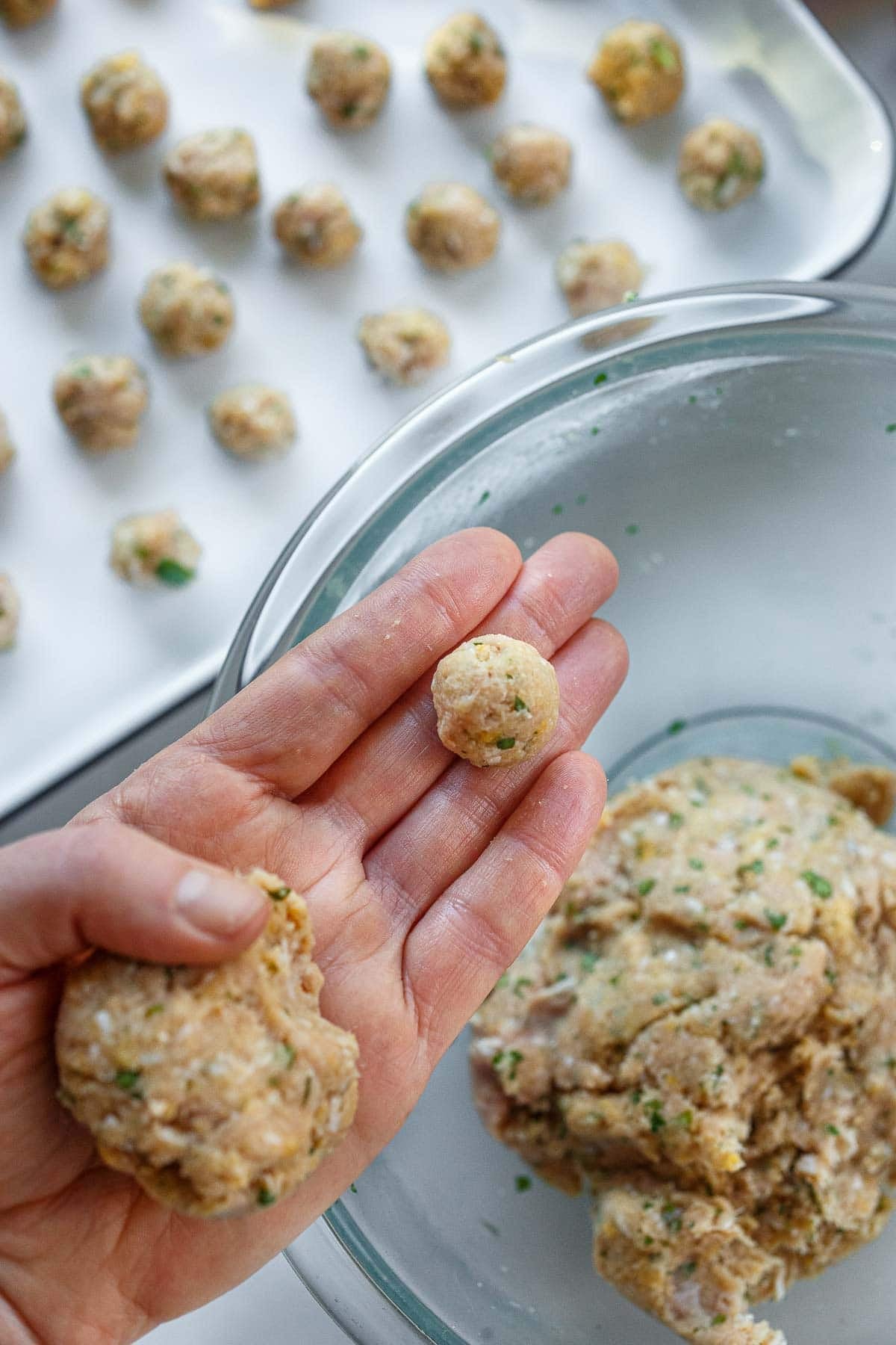 glass mixing bowl with hand held over top with handful of meatball mixture and a smaller sized meatball. 