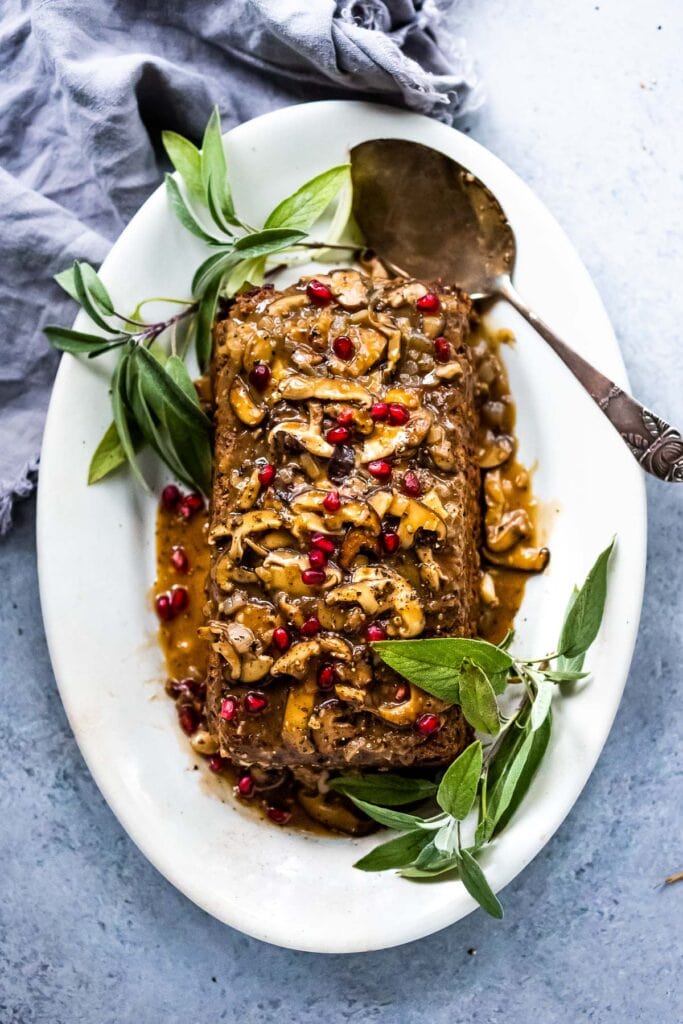mushroom loaf with mushrooms and pomegranate on top on plate with serving utensil and sage leaves. 