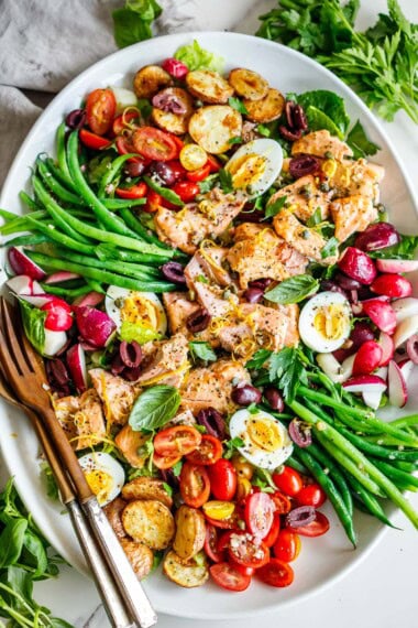 nicoise salad with salmon, boiled eggs, roasted potatoes, green beans, olives, radishes, tomatoes, and herbs on serving platter with utensil, one of our Mediterranean recipes.