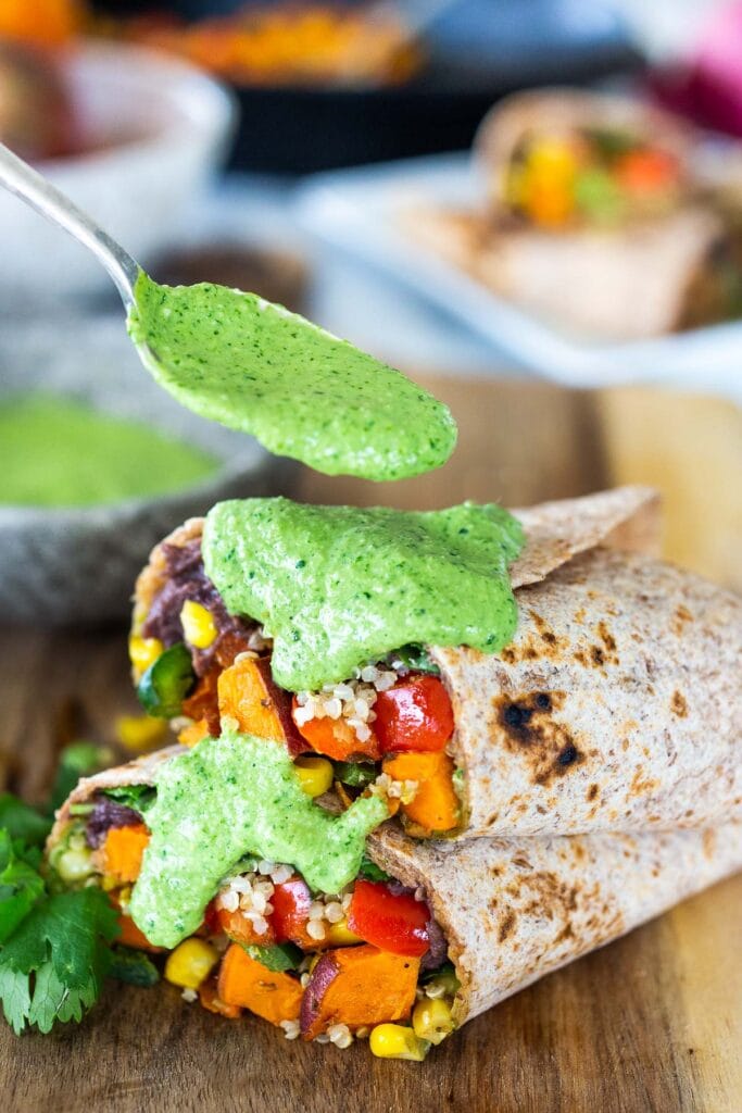 Peruvian burritos with sweet potato, creamy black beans, veggies, and quinoa with a spoon drizzling aji verde sauce over top.