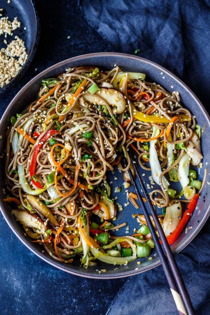 bowl of saucy sesame soba noodles with mushrooms, peppers, carrots, sesame seeds, green onions with chopsticks. 
