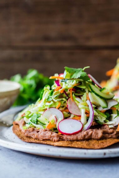 How to make Tlayudas! This healthy, lightened-up, version is vegan and GF adaptable! Feel free to add walnut chorizo,  chicken carnitas or turkey chorizo! Made with a crispy tortilla, topped with refried beans, cheese (optional) cabbage slaw, avocado and cilantro.
