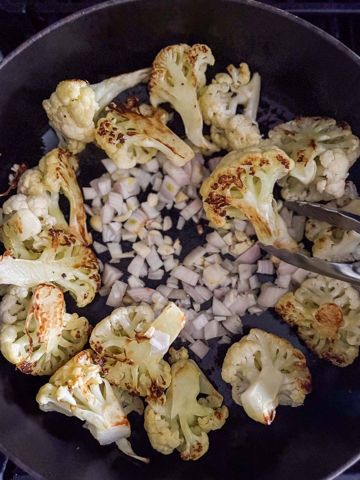tongs flipping seared cauliflower florets with chopped shallots and garlic in center of pan. 