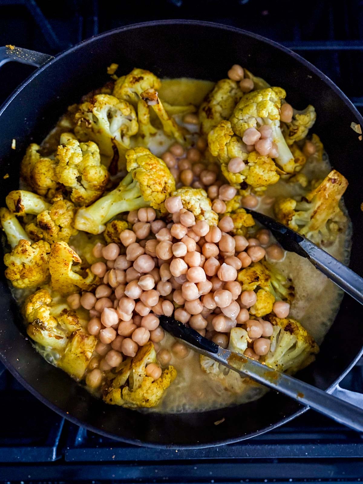 tongs stirring cauliflower curry in pot with chickpeas. 