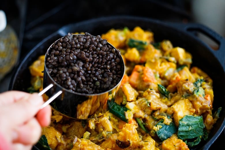 adding cup of cooked black lentils to cast iron skillet with vegetable stew base. 
